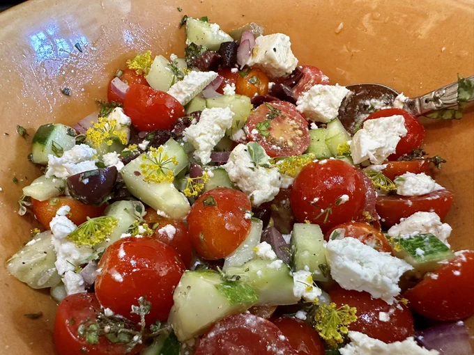 An orange bowl filled with chopped cucumbers, cherry tomato, red onion, Kalamata olives, feta and herbs.