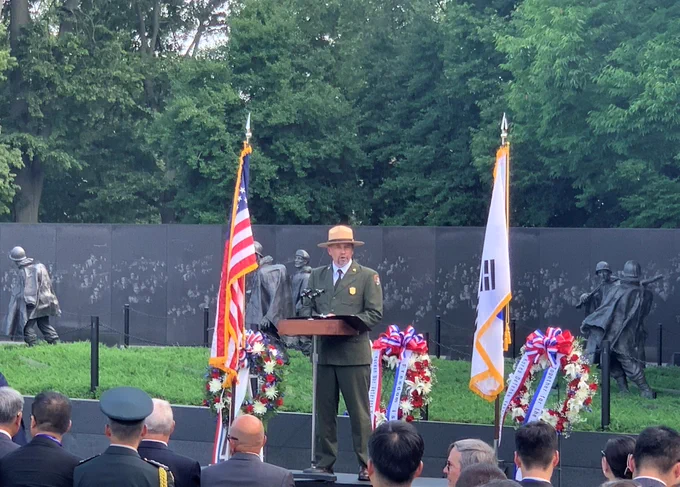 A park ranger in uniform standing between two flags and three wreaths of flowers in an outdoor military memorial.