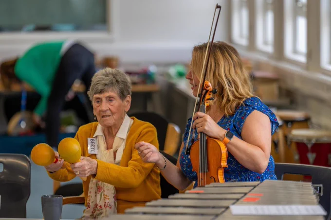 A woman with a violin gesturing towards another woman playing some maraccas.