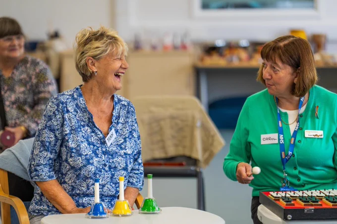 A woman with small multicoloured bells laughing next to another woman playing a xylophone.