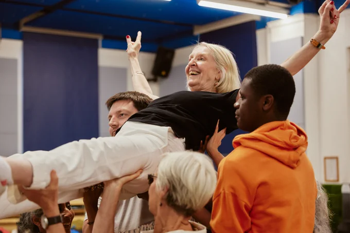 A woman smiles as she is carried by other cast members. 