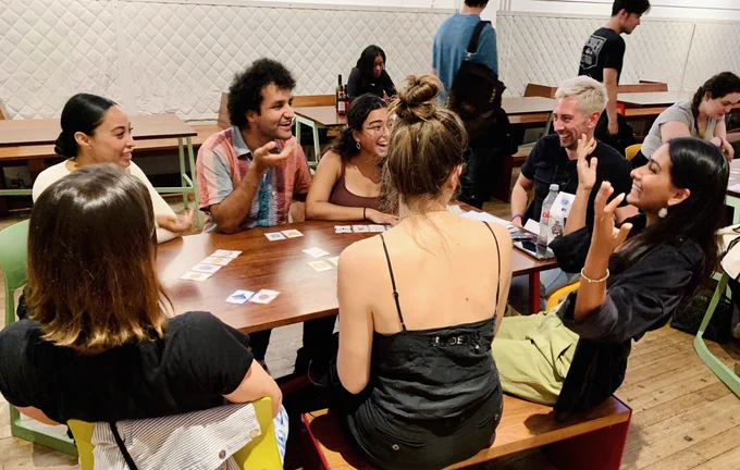 5 women and 2 men sit around a wooden table. The woman on the corner leans back and throws her hands up in a state of funny surprise. The others are looking at the woman and laughing. On the table are the Hands Off my Hippo cards, showing a number of different animal faces