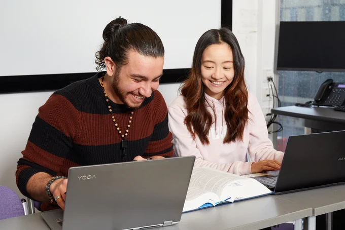 Two people laughing as they read a book and work on their laptops.
