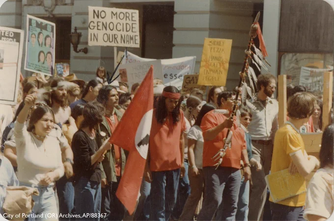 Color photograph from 1976 of a group of protestors marching down a street. A native woman on left wears a white shirt and jeans and has her right hand raised in a fist. A man in the middle has on a red shirt with a red bandana wrapped around his shoulder length hair as a headband. Members of the crowd carry a UFW AFL-CIO flag and various protest signs, the most of prominent of which reads "NO MORE GENOCIDE IN OUR NAME". Another man in a red shirt and jeans carries what appears to be a speaker's staff or spear that is adorned with feathers.