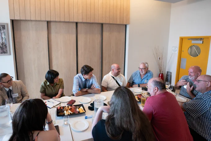 Prime Minister Justin Trudeau and Minister Ginette Petitpas-Taylor are sitting at a table with a group of people. One person is speaking and gesturing with their hands.