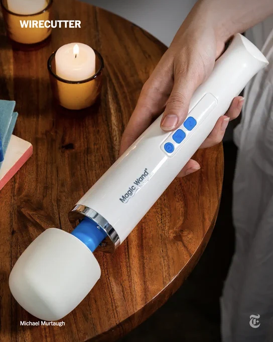 A hand holding the long, white Magic Wand vibrator over a round wooden table that has candles and books.