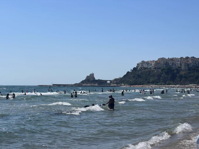 Sperlonga beach with a multitude of beach goers in the waves at the front and the castle and Torre Truglia in the background connected with a bridge to the village of Sperlonga