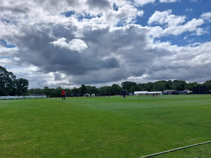 A green cricket ground with big grey clouds overhead