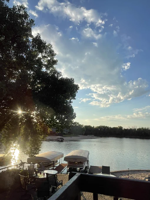 Sunset shining through trees with a lake and pontoon boats floating near the deck.
