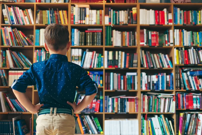 The back of a child, standing and looking at a bookcase full of books.