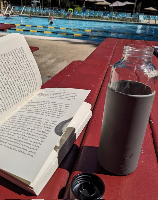 An open book and open water bottle on a picnic table with a swimming pool in the background