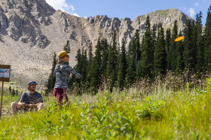 a female throws her disc from the tee box on the a-basin disc golf course, with the east wall in the background