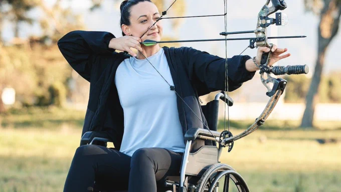 A Caucasian woman getting ready to fire a bow and arrow doing archery while sat in a wheelchair in a field on a bright day.  