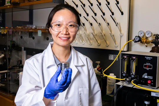 Zhen Liu (an East Asian woman in a lab coat, goggles, and gloves) holding up a small metal container with lab equipment behind her