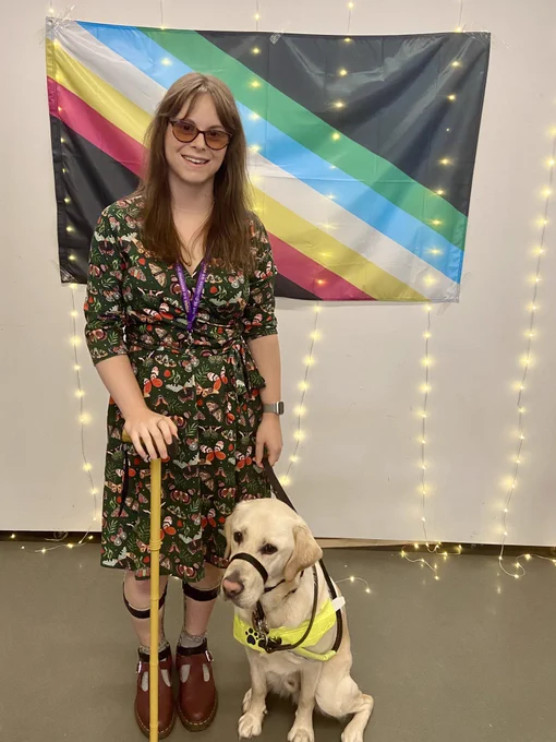 Chloe and guide dog Dezzie stood in front of a disability pride flag. Chloe is wearing AFO splints and is holding a yellow walking stick.