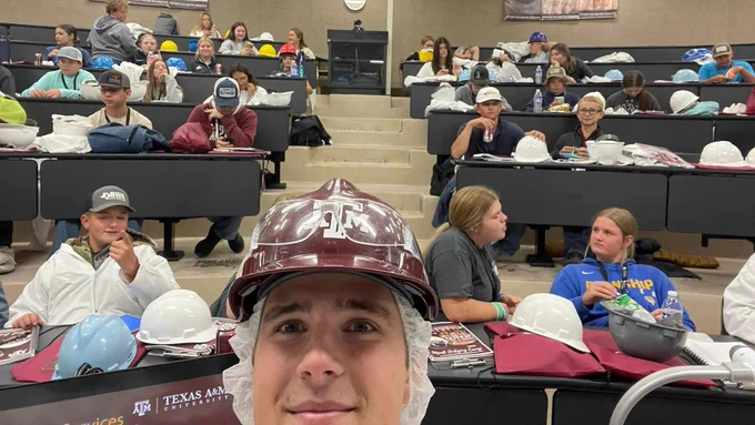 A student takes a selfie in a classroom with camp attendees behind him in classroom chairs.