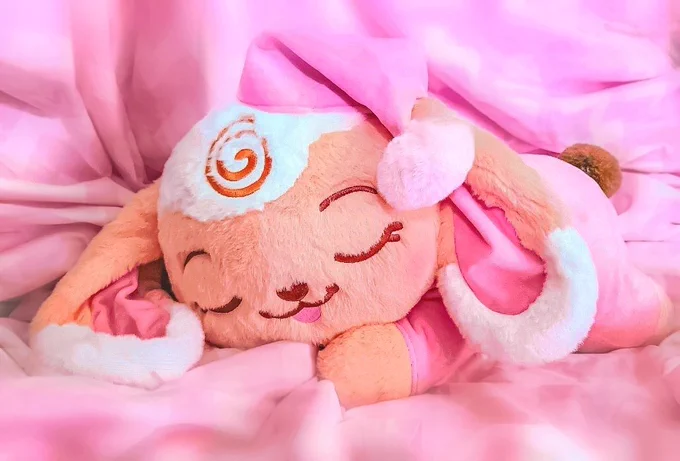 A large stylized plushie of a beige and white lop-bunny plushie wearing pink pajamas and a sleeping cap, sleeping on her stomach, her head on her paws. Background is a pink sheet.