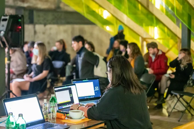 Woman working on desk with multiple laptops in an event space. Audience blurred in the background.
