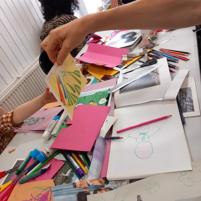 A table full of zine making materials, a hand is holding up a scrap of paper and a smaller hand is reaching for it.