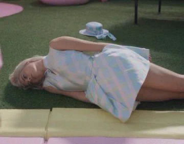 Picture of Margo Robbie as Barbie lying down on the grass in a blue plaid dress from the Barbie movie.