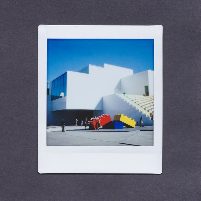 Instax print on a dark grey background, showing the LEGO House, Billund, Denmark, with giant LEGO bricks and visitors outside.