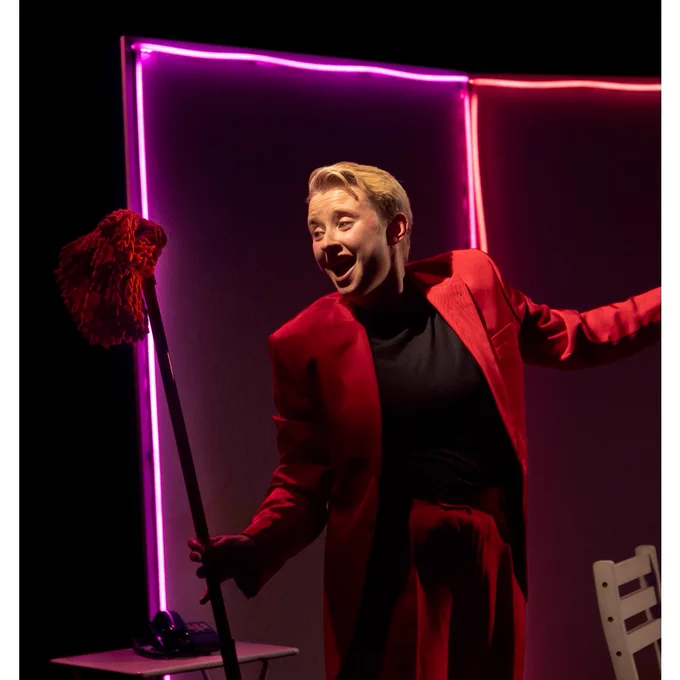 Hannah wearing a red suit, sining dancing with a red mop. Backdrop is outlined with pink and purple neon lights