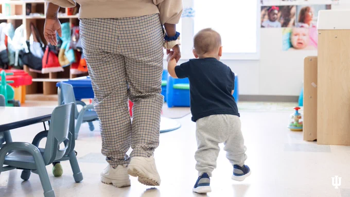 A small toddler, shown from behind, holds hands with a teacher and walks through the classroom in the Center for Young Children at IUPUI.