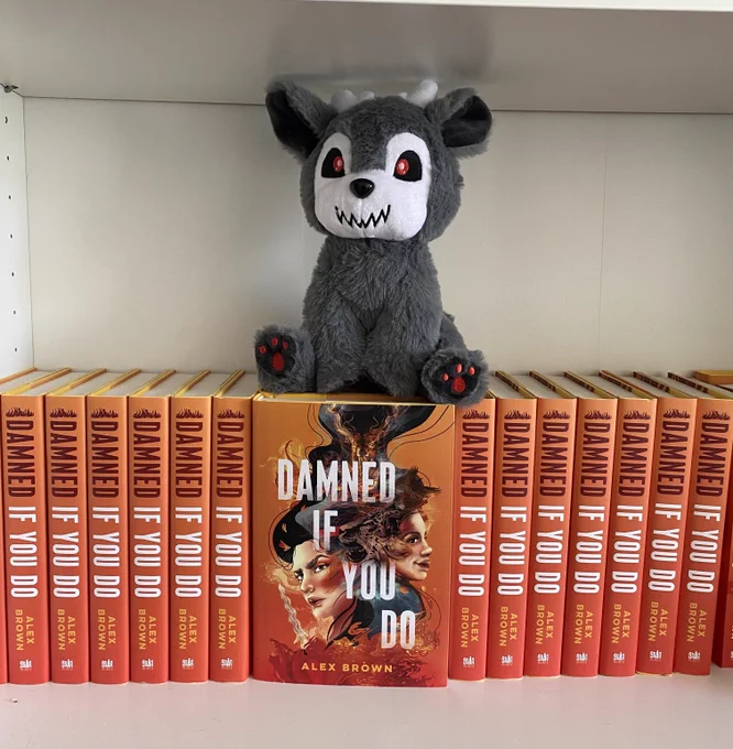 Hardcover copies of DAMNED IF YOU DO sit on a bookshelf with a cryptid plushie on top of them