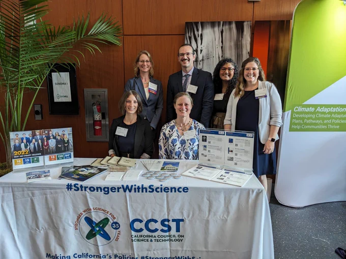 A group of CCST staff standing behind their booth with a CCST tablecloth and misc. items on the booth.