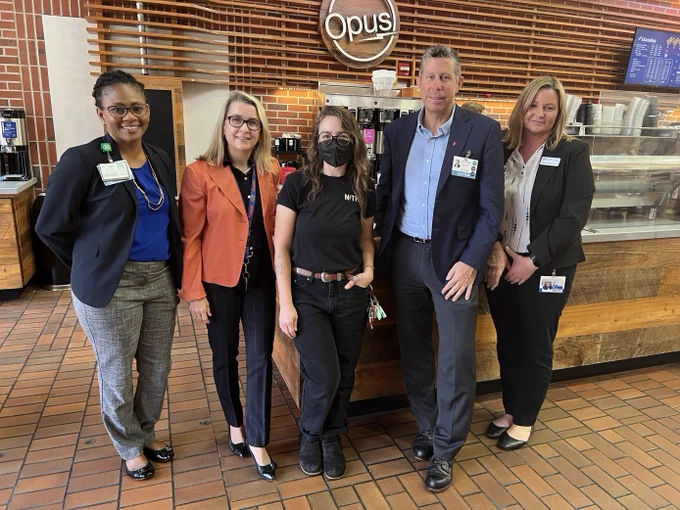 Five people standing in front of an Opus Coffee bar in the UF Health Shands Hospital Atrium. The person in the center is dressed in all black with an all black face mask on.