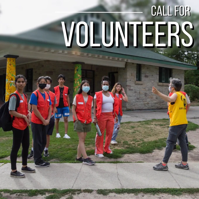 Photo of a group of 7 volunteers in bright orange vests. They gaze off to the side as a person in a yellow vest extends their arm in the same direction.