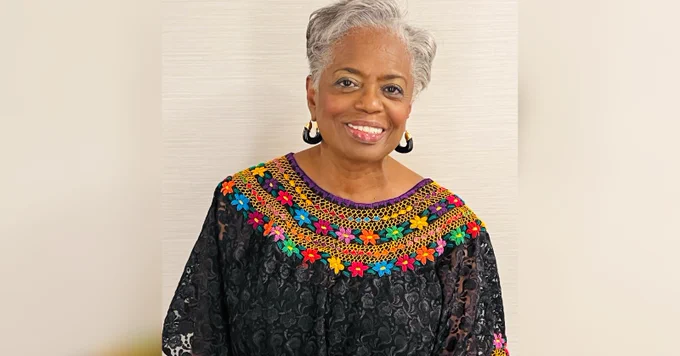 Headshot of Janice Welburn. She stands in front of a white wall, wearing a black floral top with a colorful, floral patterned neckline, and black hoop earrings with gold ends. She has white, short hair, and a smile on her face.