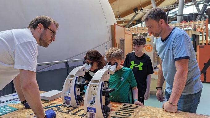 Two members of a family stare down microscopes while two more family members look on. A Science Explainer is also there to help. 