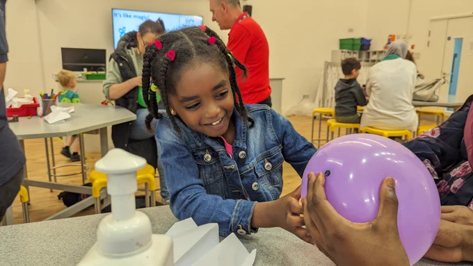 A young girl is touching a purple balloon and smiling. 