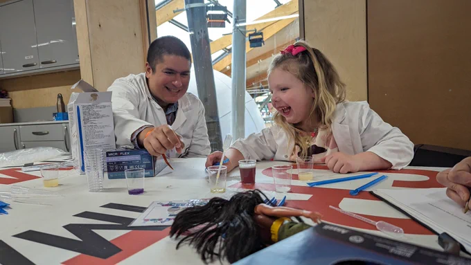 A young girl and a Science Explainer, both wearing lab coats, laugh and reach for a pipette. The table has small glasses with different coloured liquids in them. 