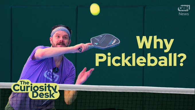 A white man in a purple T-shirt reaches with a pickleball paddle for an incoming yellow ball. The wall behind him is green and on screen it says "why pickleball" with yellow writing. 
