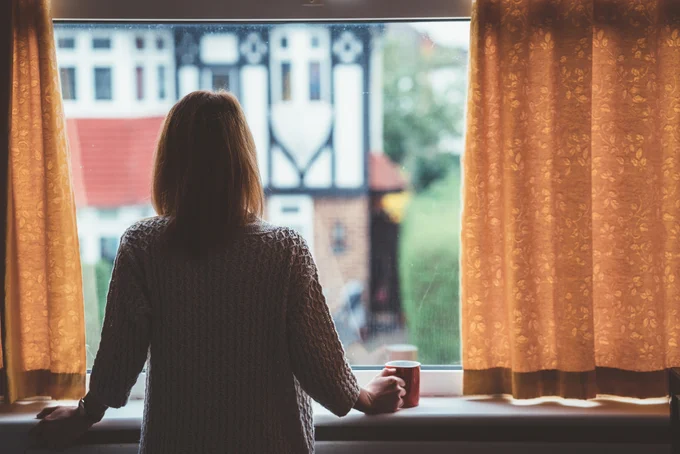 A woman with short hear looks out of her window holding a coffee mug