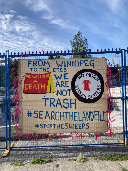 Image ID1. A large banner affixed to a blue rental fence. Text: “From Winnipeg to the DTES, we are not trash. #SearchTheLandfill #StopTheSweeps. A red and yellow tent is depicted on the left with the text “displacement = death” and a black and white emblem with a red dress in the centre is depicted on the right, with the text “No more stolen sisters.” The banner is surrounded by red ribbons.