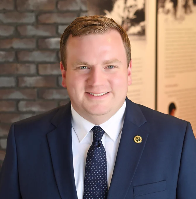 Photo of Brian Allman wearing a navy blue suit with a white shirt and navy blue tie with white and black cubes on it in front of a brick wall.