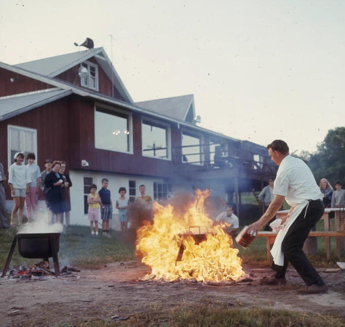 A historical photo of a person tending to a fish boil, which is a big ball of fire, as spectators look on.