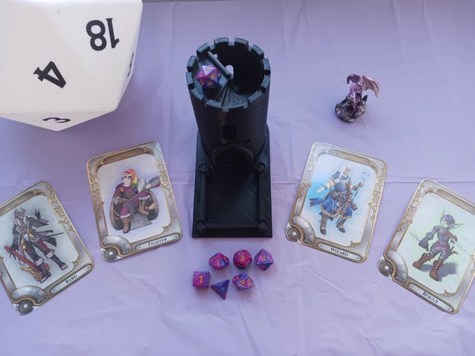 A purple tablecloth.
On the table there is a black dice tower, a set of purple polyhedral dice for Dungeons and Dragons, a purple dragon miniature figurine, and a large white 20-sided dice sculpture.
There are also cards with four characters on them.
A bard in black clothes with an electric guitar.
A fighter with rainbow hair, a big axe, and a battle wheelchair.
A wizard with blue robes and a staff,
A green Goblin rogue with purple hair and dual daggers.