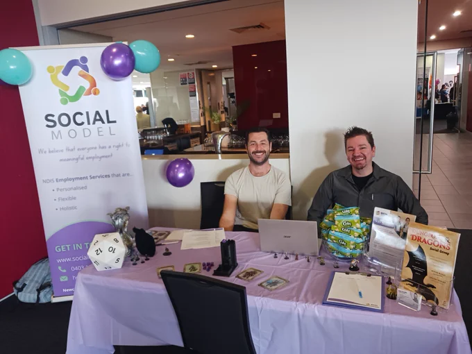 two smiling men sitting behind a brightly coloured  purple expo stall.
There are balloons, flyers, plus Dungeons & Dragons decorations.