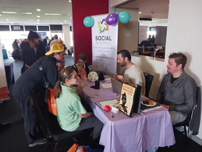two men sitting behind a brightly coloured and decorated purple expo stall.
They are running a game of dungeons and dragons for a group of four high school students.