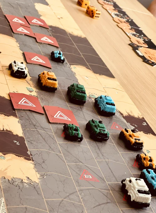 A map of a road split into chevron-shape spaces. Cars in 4 different colours (white, orange, blue, green) are all placed on different spaces facing away from the picture. Red cardboard chevrons cover some of the spaces, with hazard triangles on top