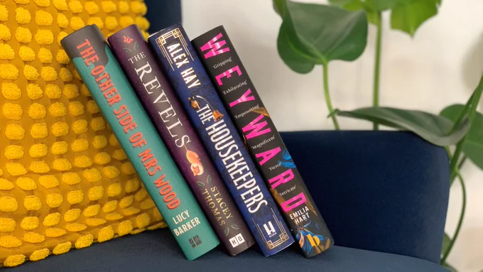 Four hardback books stacked vertically on a blue chair. The books are The Other Side of Mrs Wood by Lucy Barker; The Revels by Stacey Thomas; The Housekeepers by Alex Hay and Weyward by Emilia Hart. 