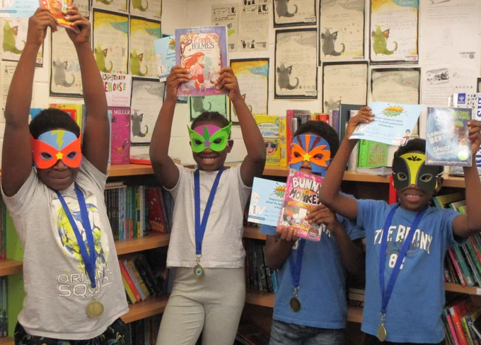 four children wearing masks holding books aloft