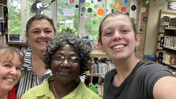 Four women smile at the camera. The one on the right is holding the camera, as if to take a selfie. They are taking the photo in a library.