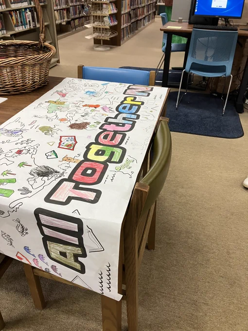 A large banner spread out on a table reads "All Together Now". The banner looks like it was printed out in black and white and, over a long period of time, people have colored in various pictures and words on the banner.