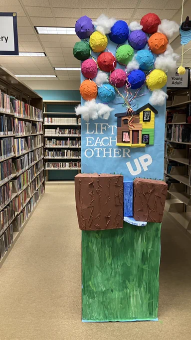The endcap display of a library shelf. The display is of a house being lifted up by a dozen or so balloons into the sky above a waterfall. This is obviously the house from the Pixar movie 'Up', as shown by the caption on the display: "Lift Each Other UP."