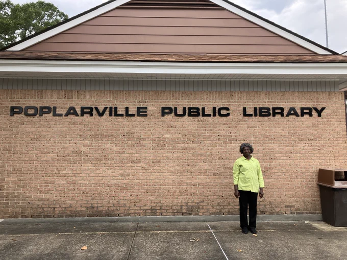 Ruth, a Black woman wearing a green shirt, stands in front of a brick building. The sign on the wall of the building says Poplarville Public Library.